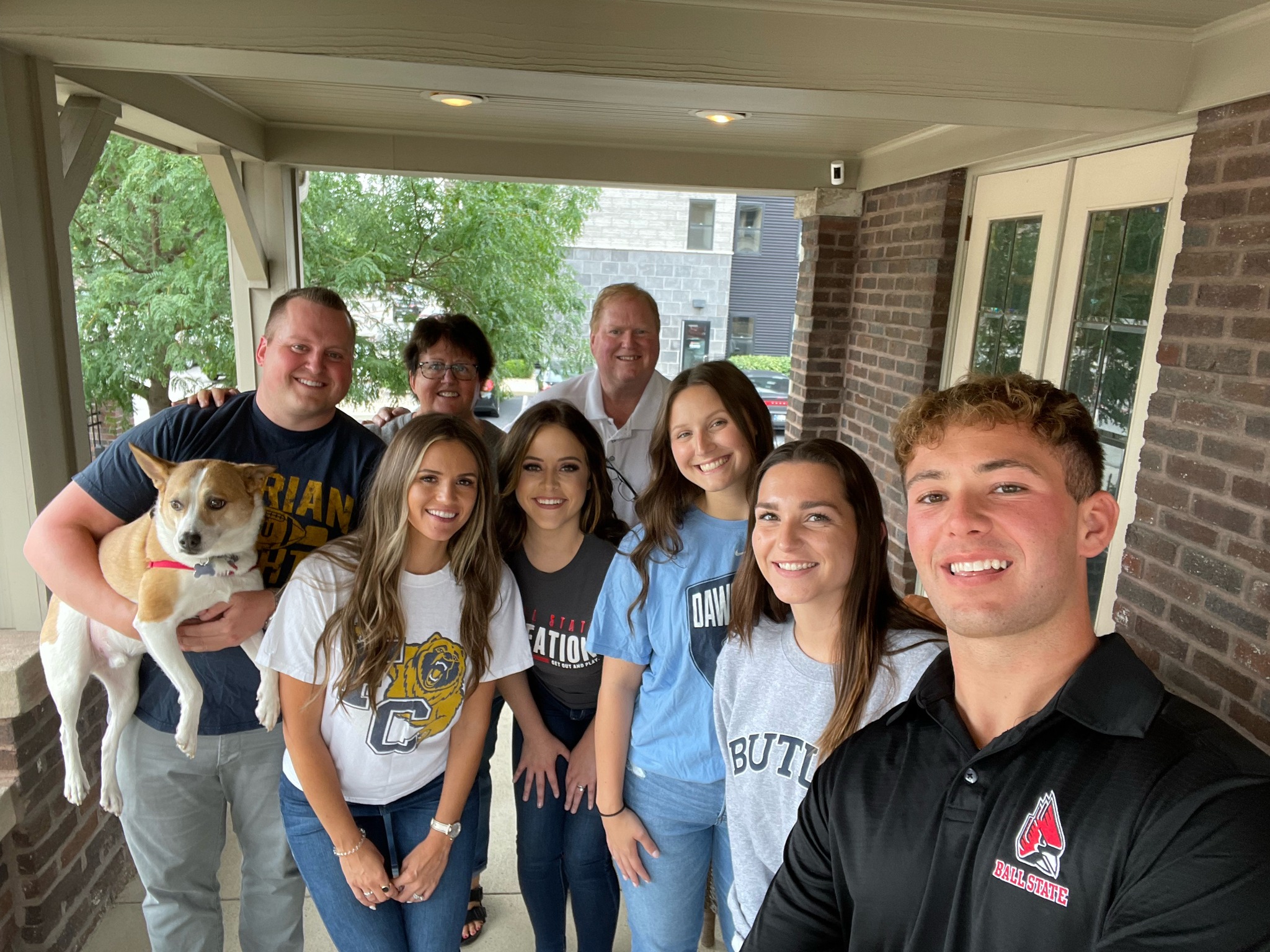 A cheerful group of eight with a dog, wearing collegiate apparel, posing for a selfie on a porch
